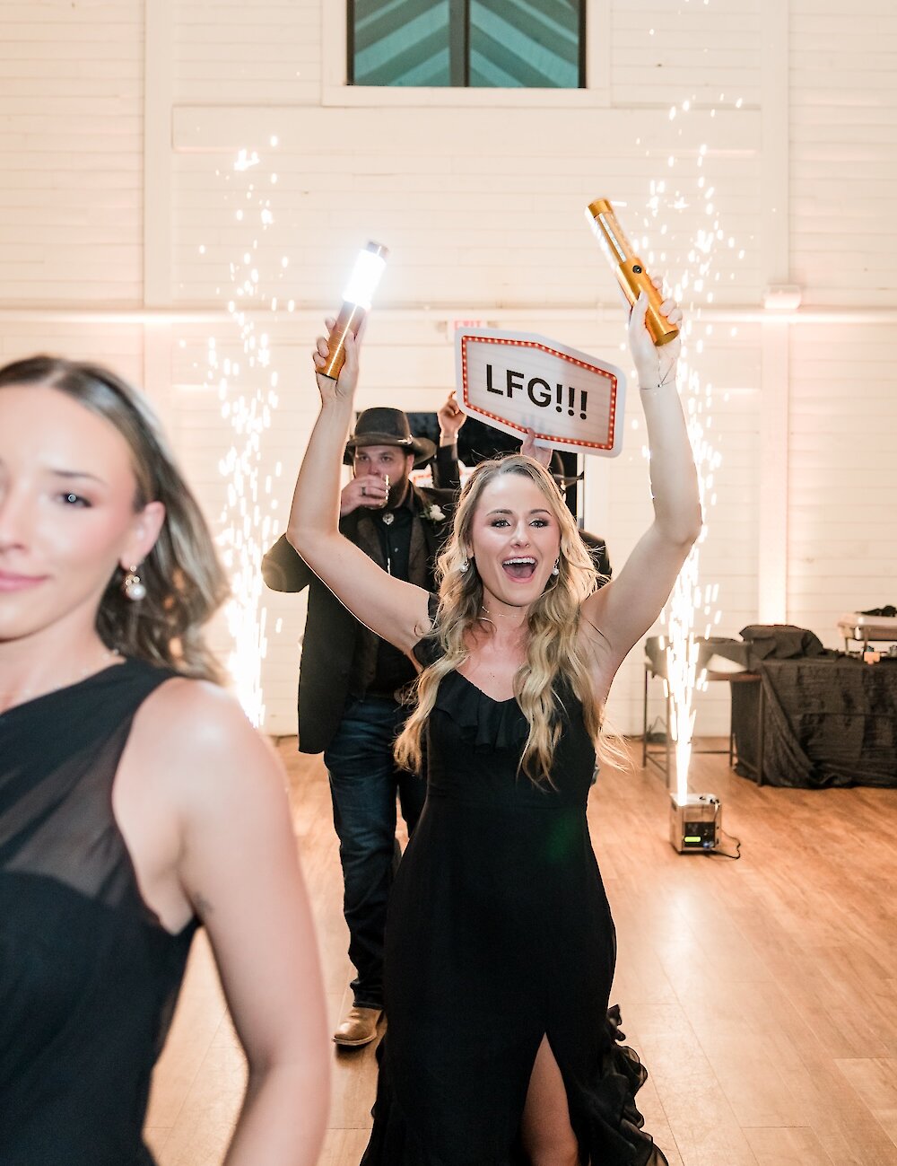 A joyful guest raises her hands holding glow sticks and an illuminated 'LFG!!!' sign, walking through a grand entrance framed by indoor pyrotechnics during the wedding reception.