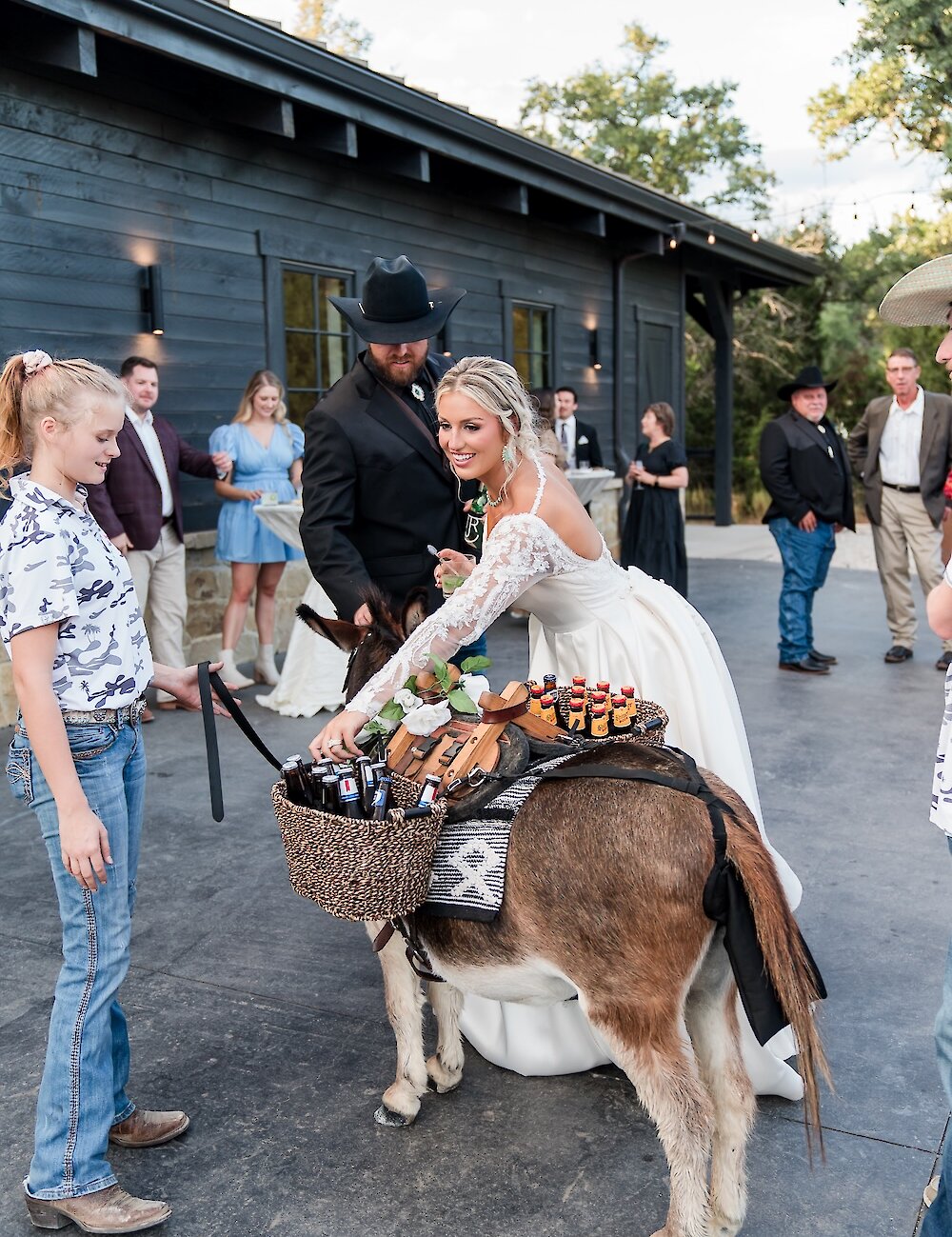 The bride and groom smile while interacting with a mini donkey carrying basket panniers of drinks and snacks for cocktail hour on the patio of a black barn venue.
