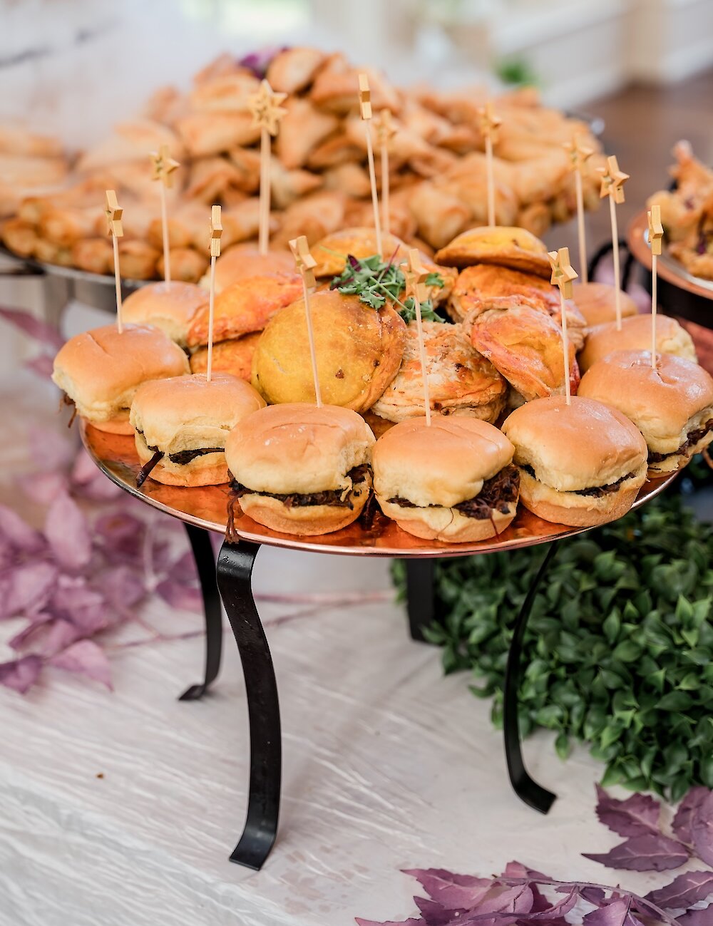 A tiered catering display holding savory food, including small beef sliders and fried pastries like samosas, served on a copper platter with star-topped toothpicks.