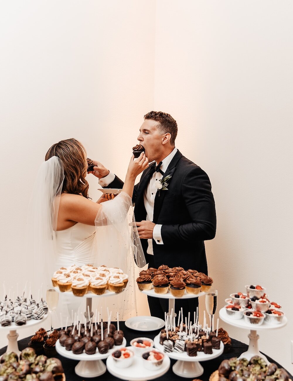 The bride and groom feed each other chocolate cupcakes from a decadent dessert table laden with various miniature sweets on tiered stands.