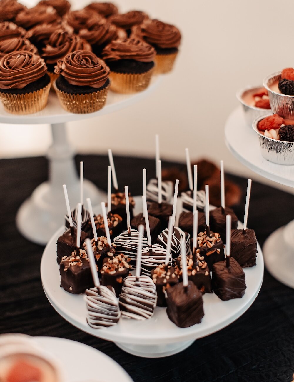 A close-up of a dessert table featuring chocolate-dipped cake pops on sticks, mini chocolate cupcakes, and fruit tarts on white tiered stands.