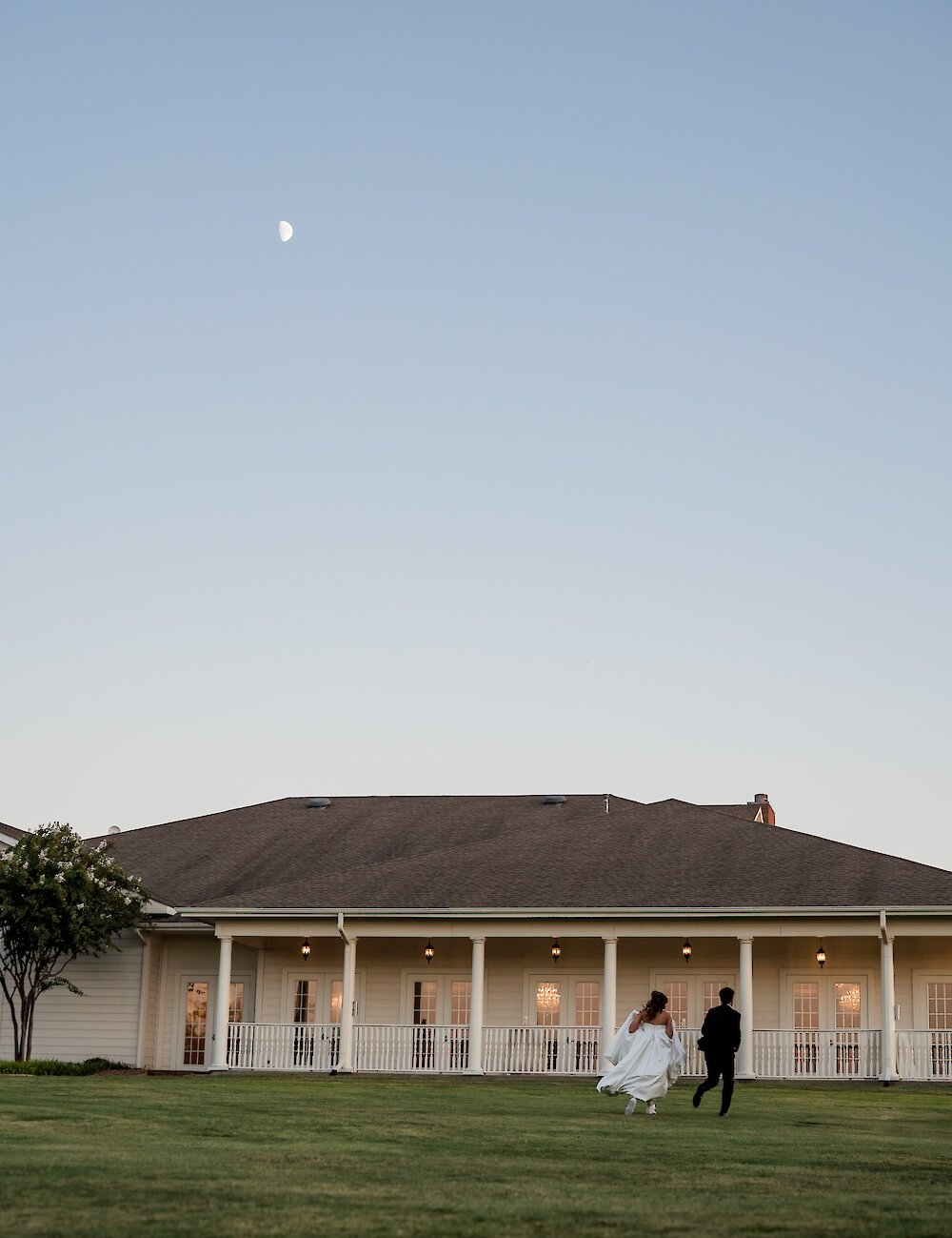 The bride and groom run across a wide, grassy lawn toward a large white southern-style mansion at twilight, with the crescent moon visible in the sky.