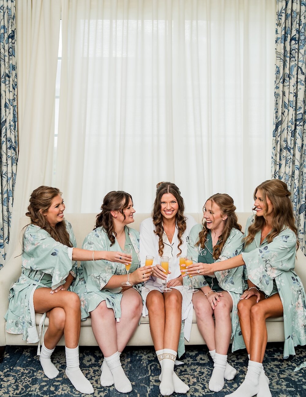 The bride and four bridesmaids, wearing coordinating robes and socks, sit on a couch raising champagne glasses for a joyful portrait.