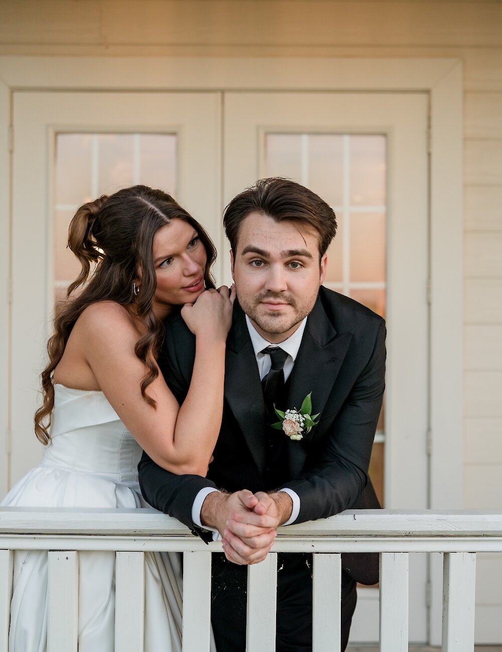 The bride lovingly leans on the groom, who is looking straight ahead, while posing on a white wooden porch railing at sunset.