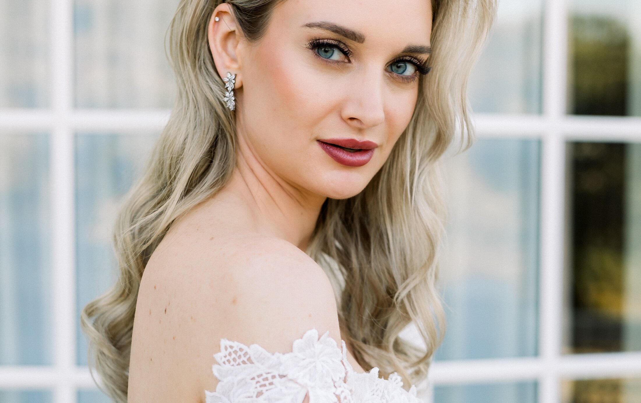 Close-up portrait of the beautiful bride in a lace off-the-shoulder gown, wearing diamond earrings and ring, posing in front of a glass window.