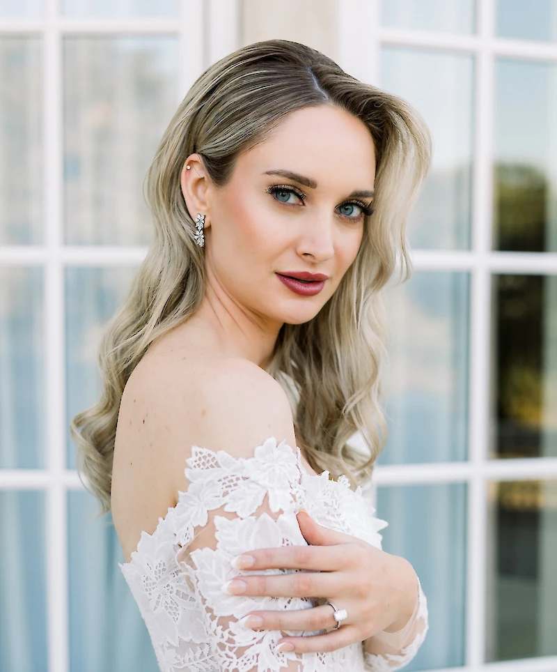 Close-up portrait of the beautiful bride in a lace off-the-shoulder gown, wearing diamond earrings and ring, posing in front of a glass window.