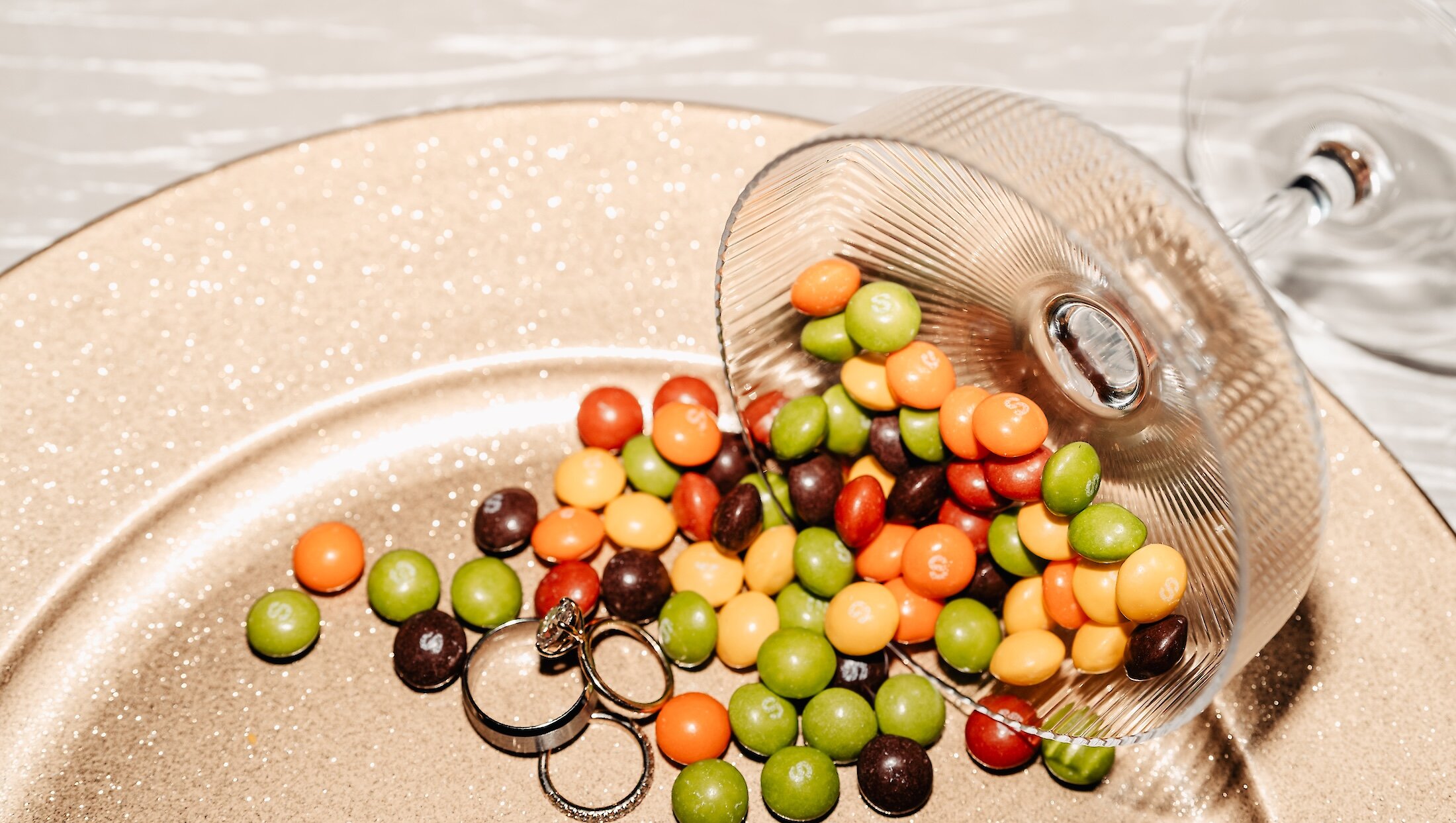 The wedding bands and engagement ring are photographed next to a pile of spilled Skittles candy on a gold glitter charger plate.