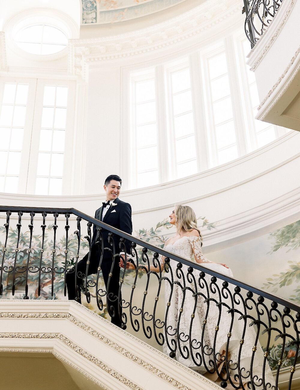 The bride and groom are holding hands and smiling at each other while walking up a grand ornate black wrought-iron staircase in a brightly lit atrium with painted murals.