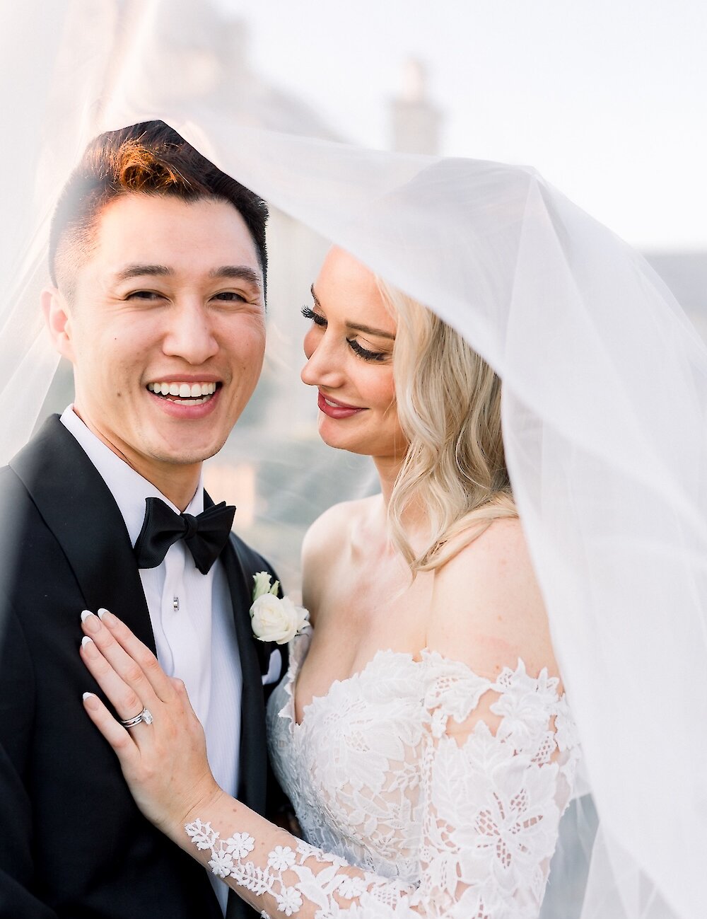 A close-up of the happy couple smiling beneath the bride's sheer veil, with the groom in a tuxedo and the bride in a lace off-the-shoulder gown, displaying her engagement ring.