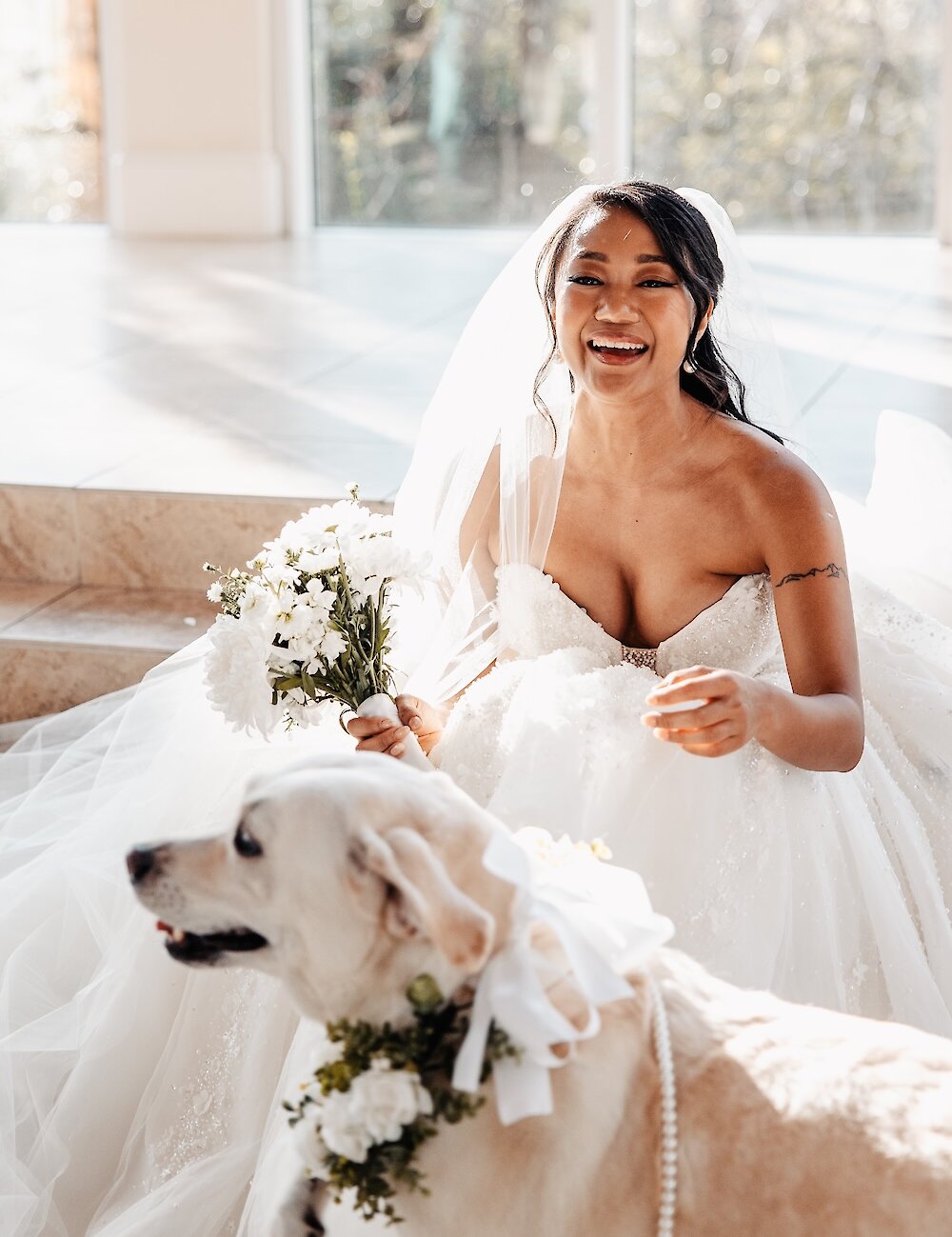 The joyful bride in a ballgown sits with her labrador dog, who wears a floral collar and pearl necklace, holding a white bouquet in a sunny room.