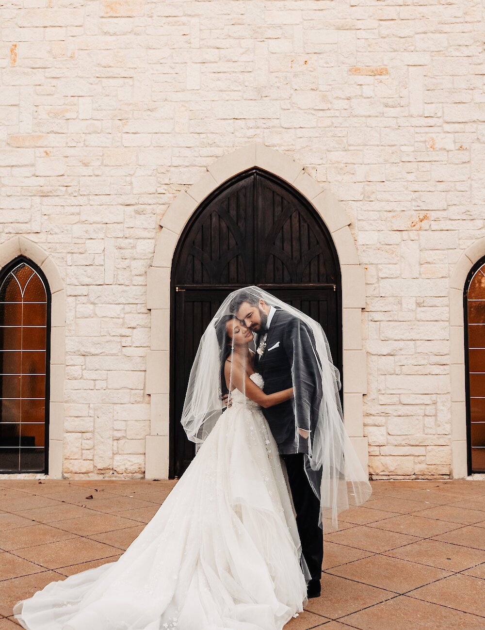 The bride and groom kiss intimately under the bride's flowing veil in front of a white stone chapel wall and a dark arched wooden door.