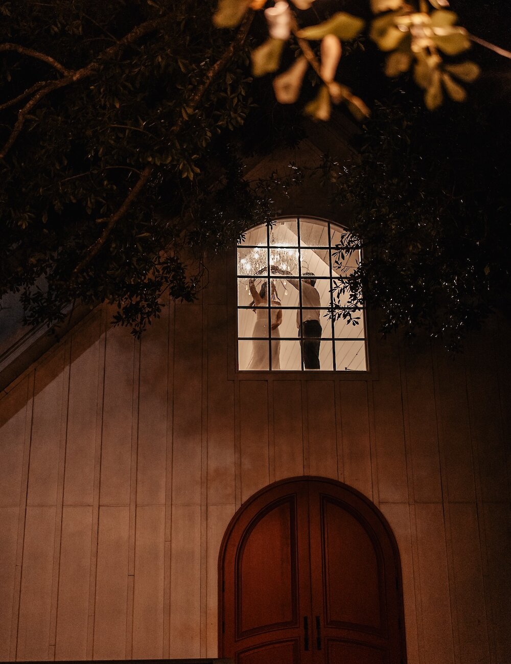 A nighttime exterior view of a chapel showing the silhouette of the bride and groom kissing in a large upper window, illuminated by a chandelier.