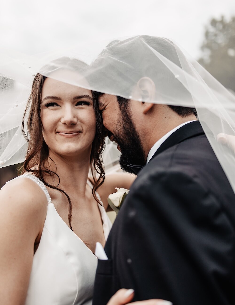 A romantic close-up of the bride smiling while the groom whispers to her, with both faces partially covered by the sheer bridal veil.