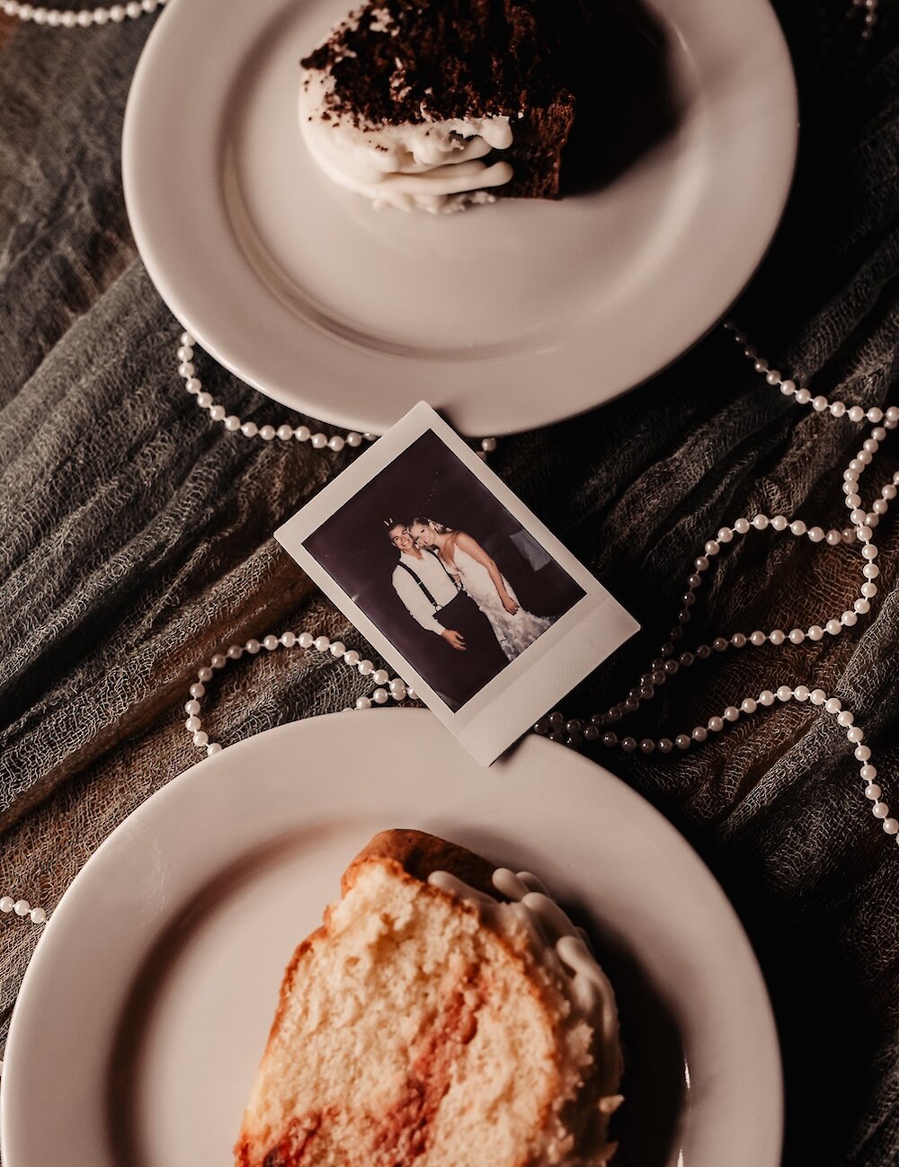 An overhead shot of two plates holding slices of wedding cake, separated by a Polaroid photo of the bride and groom, and draped with white pearl necklaces.