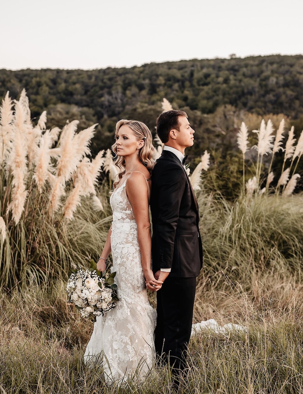 The bride and groom stand back-to-back holding hands in a grassy field with tall pampas grass, with the bride holding a white and green bouquet.