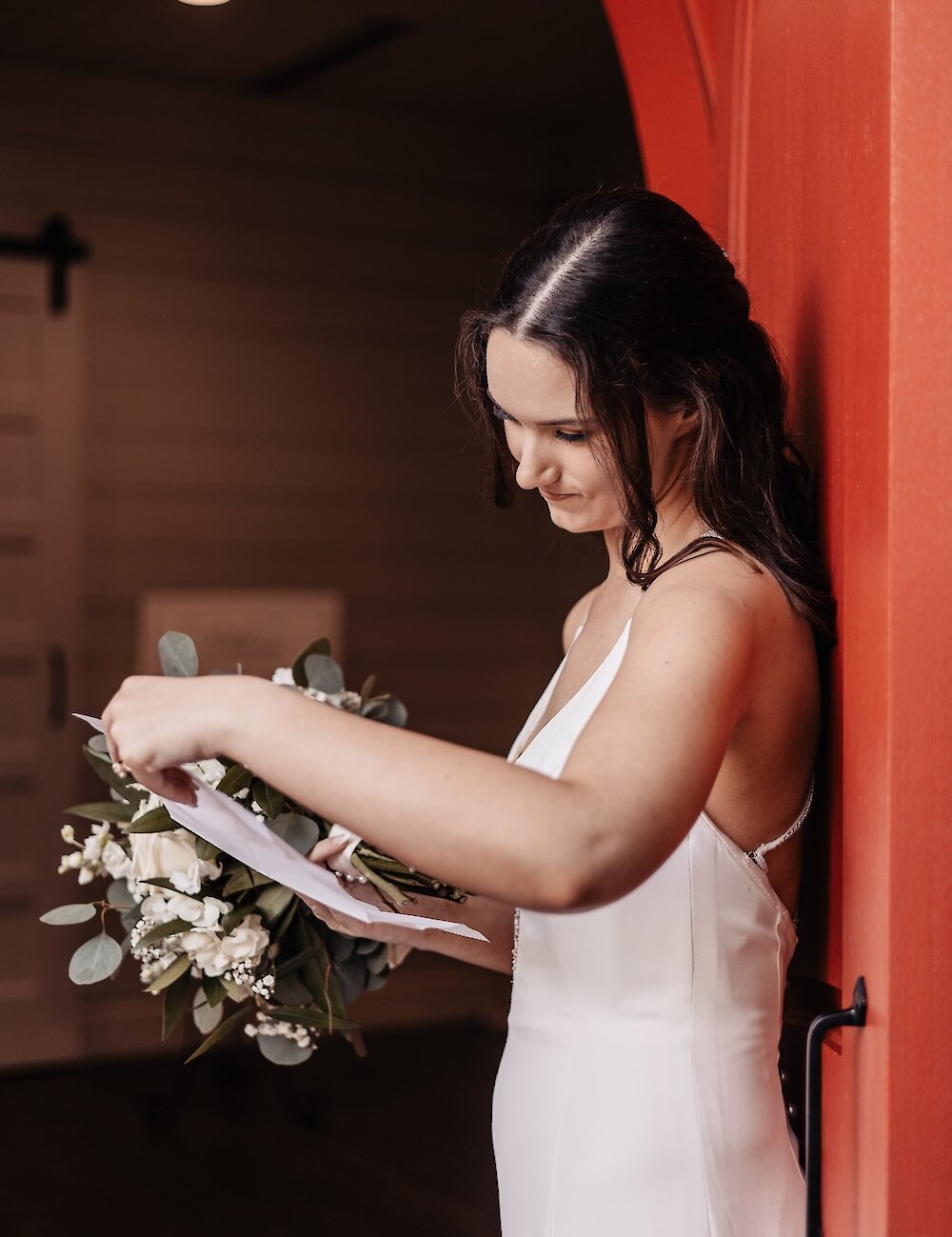 The bride in a simple white slip dress stands in a doorway, holding her white and green bouquet while reading a letter, with the surrounding frame painted bright orange-red.
