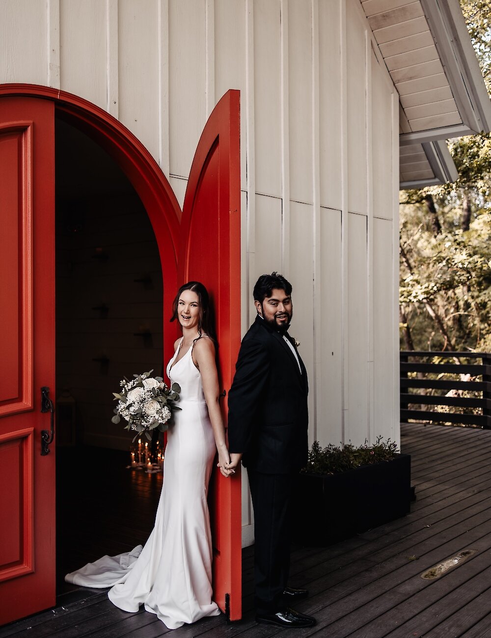 The smiling bride and groom stand back-to-back, holding hands and leaning against a large arched red door on a dark wood porch, with the bride holding a white bouquet.