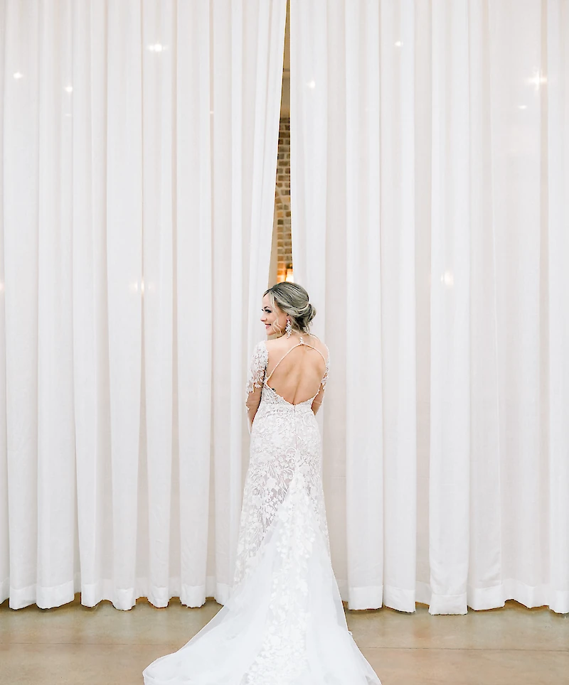 The bride stands with her back mostly toward the camera, showcasing her open-back lace gown and elegant updo against a backdrop of flowing white drapes.