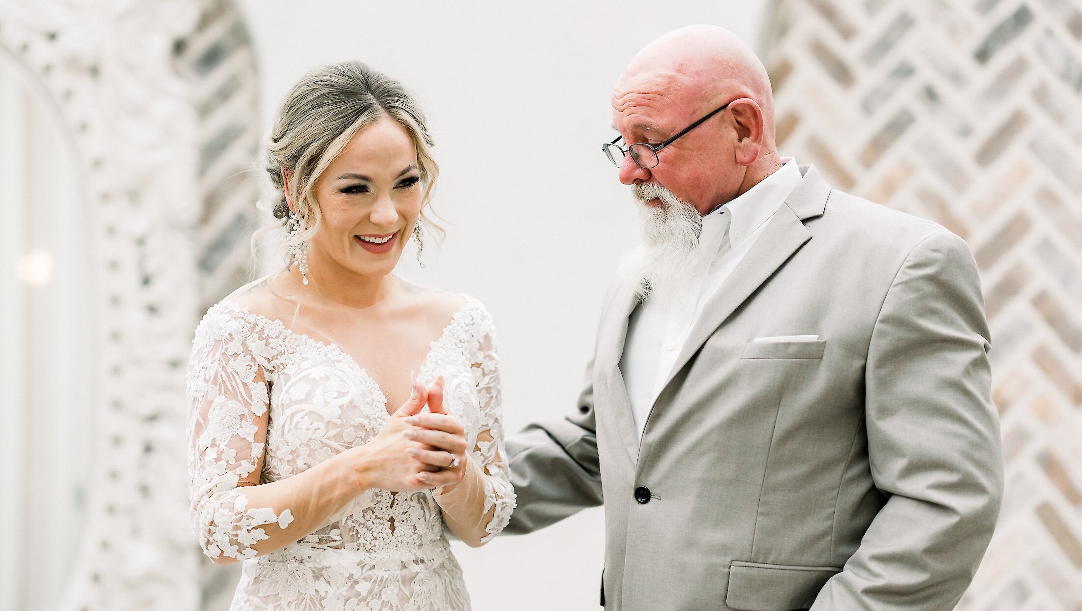 The smiling bride in a long-sleeve lace gown stands next to her father, who is wearing a light gray suit and a full white beard, during the first look.