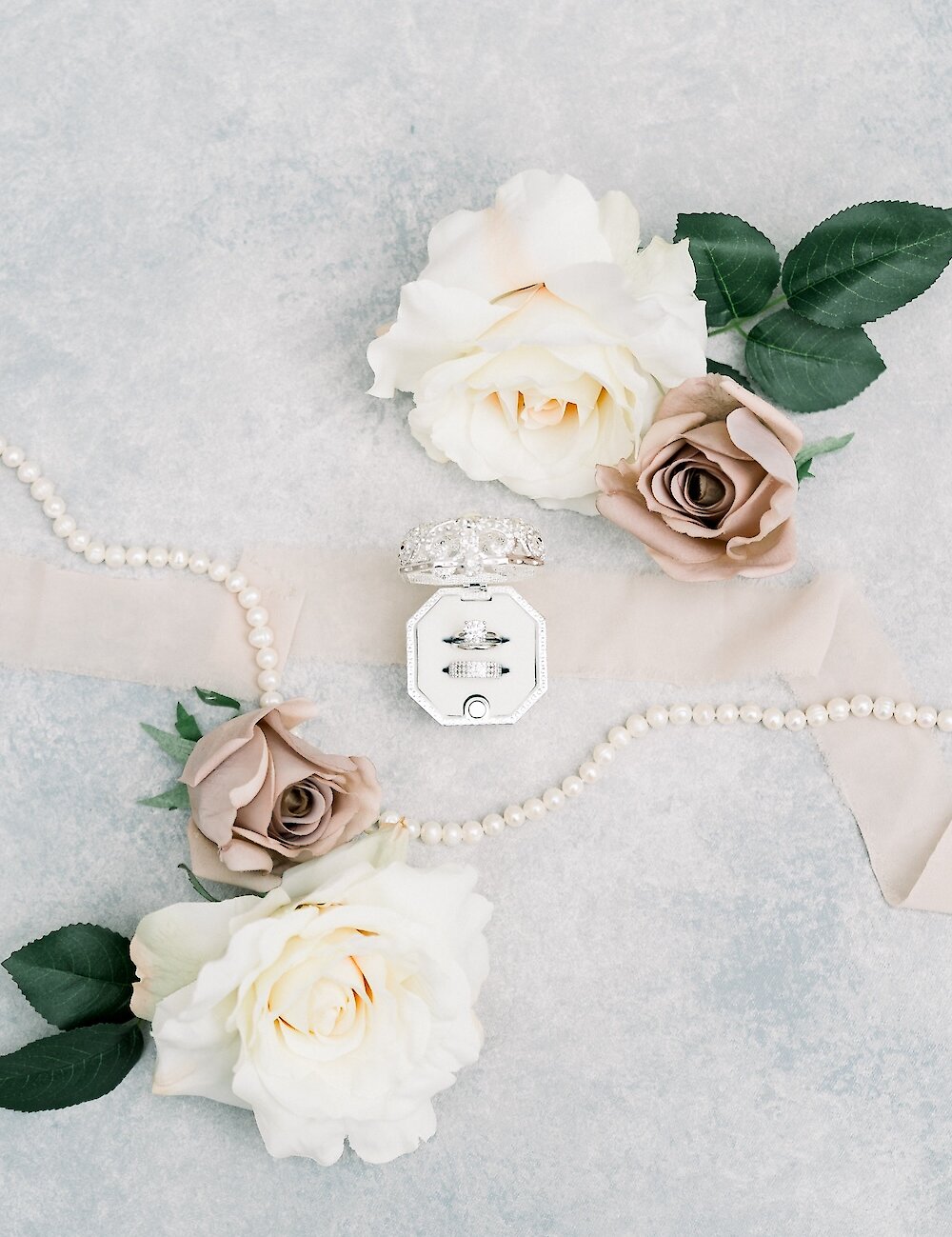 A wedding ring flat lay featuring the engagement ring and wedding bands in an octagonal box, surrounded by pearls, silk ribbon, and white and mauve roses.