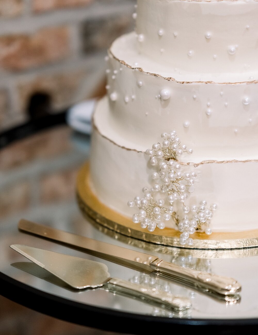 Close-up of a three-tier white wedding cake decorated with gold trim and edible pearls, next to gold serving knife and spatula on a mirrored table.