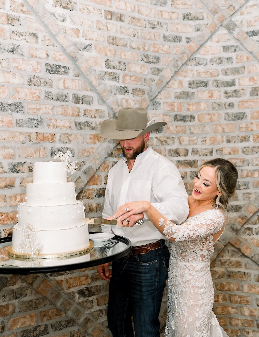 The bride in a lace gown smiles as she helps her groom, who is wearing a cowboy hat, cut their three-tier white wedding cake in front of a rustic brick wall.