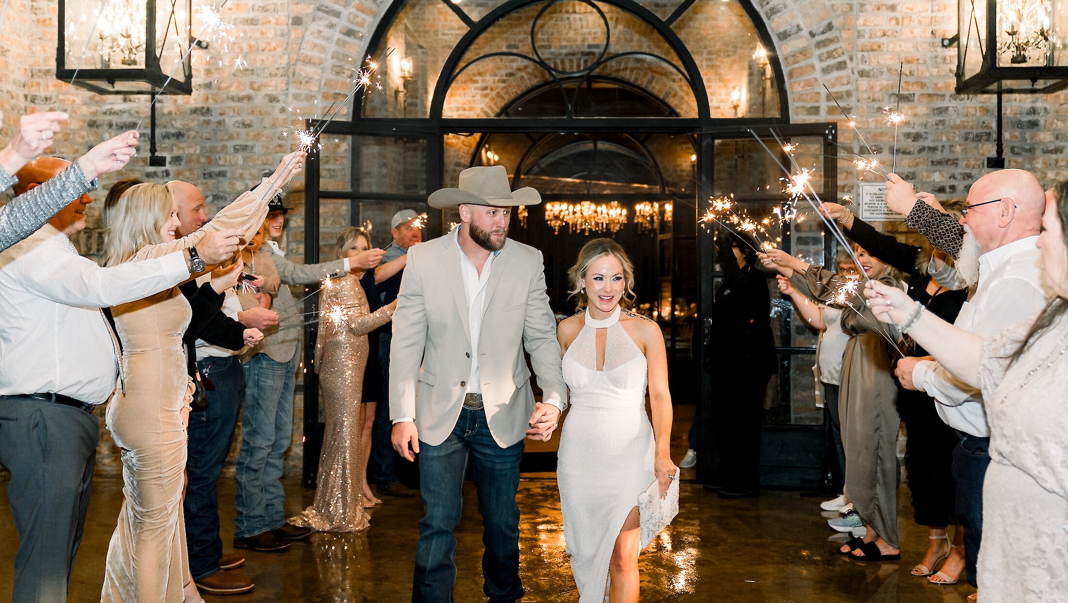 The bride and groom hold hands and smile while exiting through a tunnel of guests holding sparkling sparklers in a brick-walled venue.