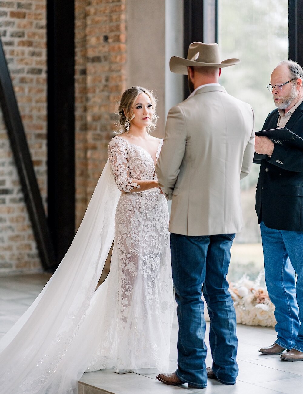 The bride in a long-sleeve lace gown looks at the groom in a cowboy hat and jeans during their wedding ceremony in front of an officiant and a brick wall.