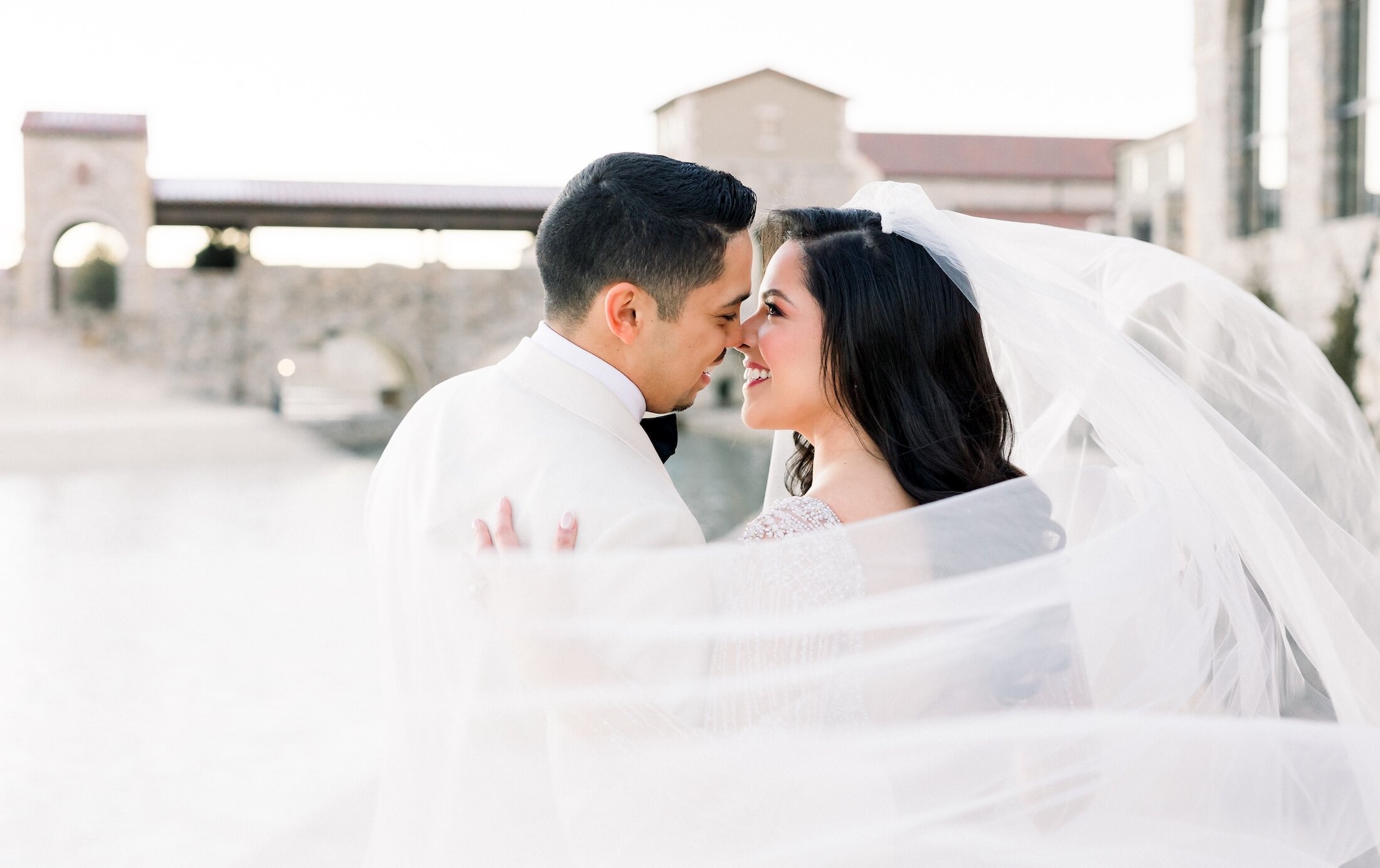 The bride and groom lean in to kiss, framed by the bride's flowing veil, with an Italian-style stone bridge and archway visible in the background.