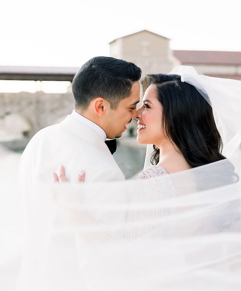 The bride and groom lean in to kiss, framed by the bride's flowing veil, with an Italian-style stone bridge and archway visible in the background.