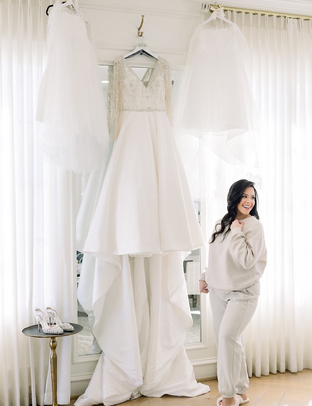 The smiling bride in a cream loungewear set stands next to her elaborate, long-sleeved wedding dress and veil hanging in the bridal suite.