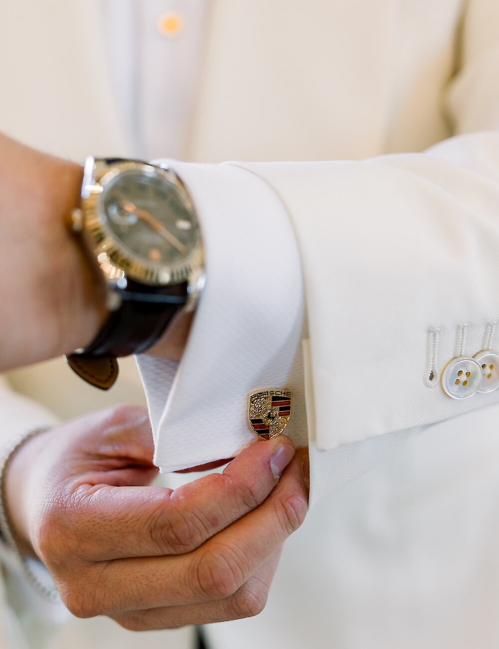 Close-up of the groom adjusting a gold Porsche logo cufflink on his white tuxedo jacket sleeve, featuring a wristwatch and button detailing.
