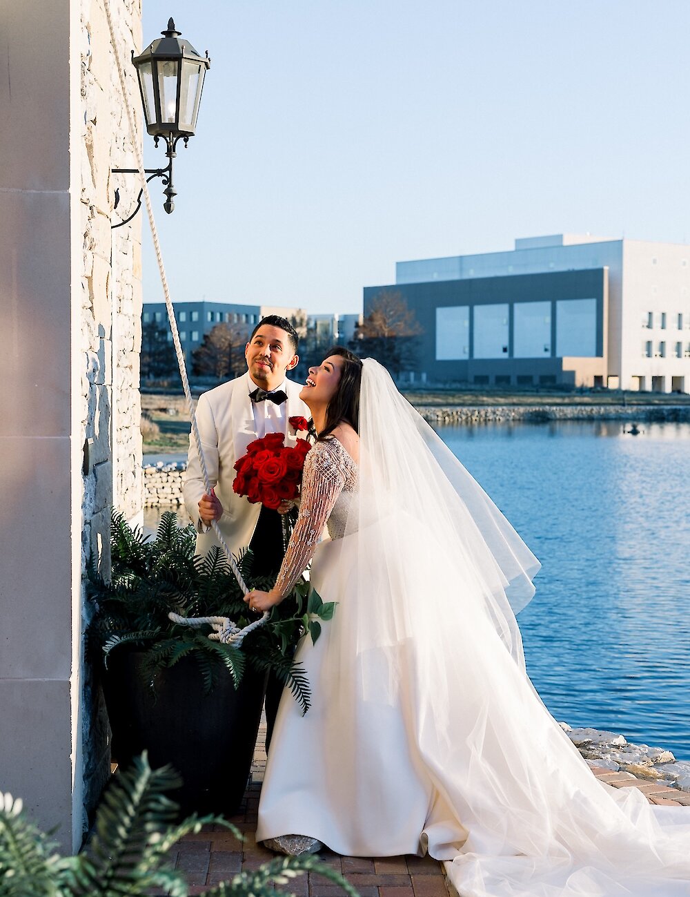 The bride and groom are smiling while pulling a rope to ring a bell on a stone wall, standing next to a planter overlooking a calm lake.