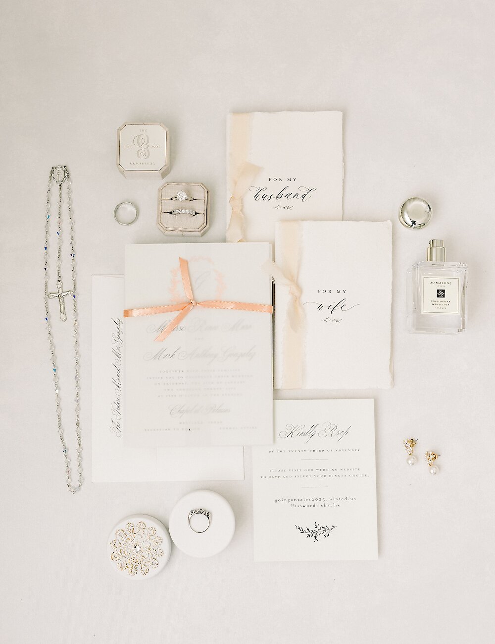 A wedding flat lay featuring the invitation suite, rings, pearl earrings, a rosary, and Jo Malone perfume bottle on a pale white background.