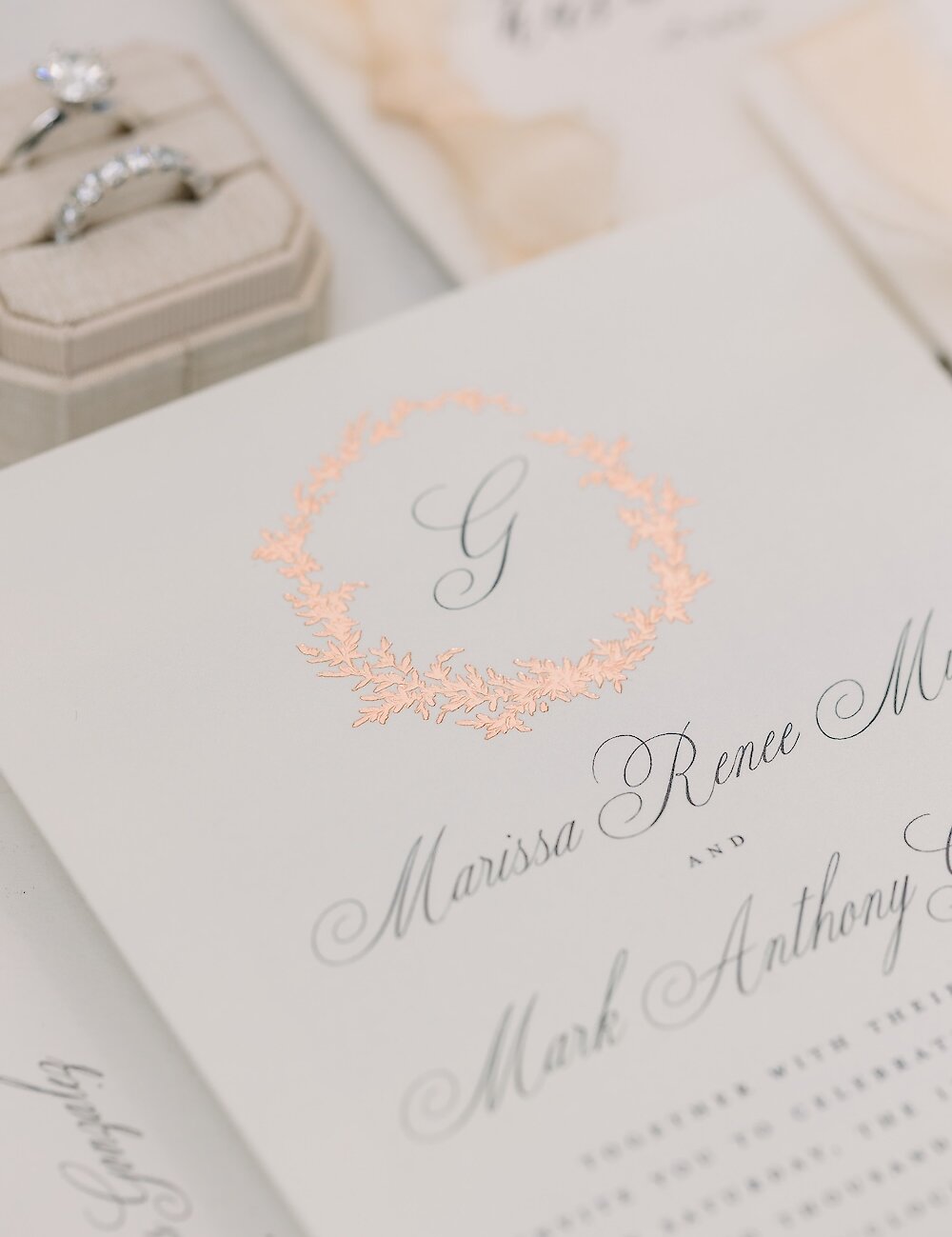 Close-up of a wedding invitation showcasing the names Marissa and Mark, with a peach floral wreath monogram detail at the top.