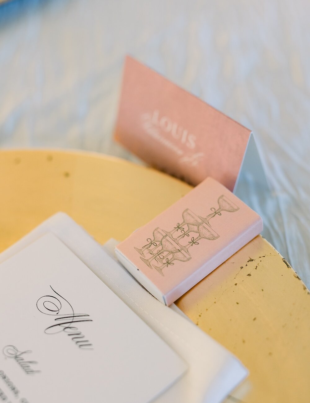 A close-up of a wedding reception setting showing a menu card, a peach place card, and a matchbox favor featuring a champagne tower illustration.