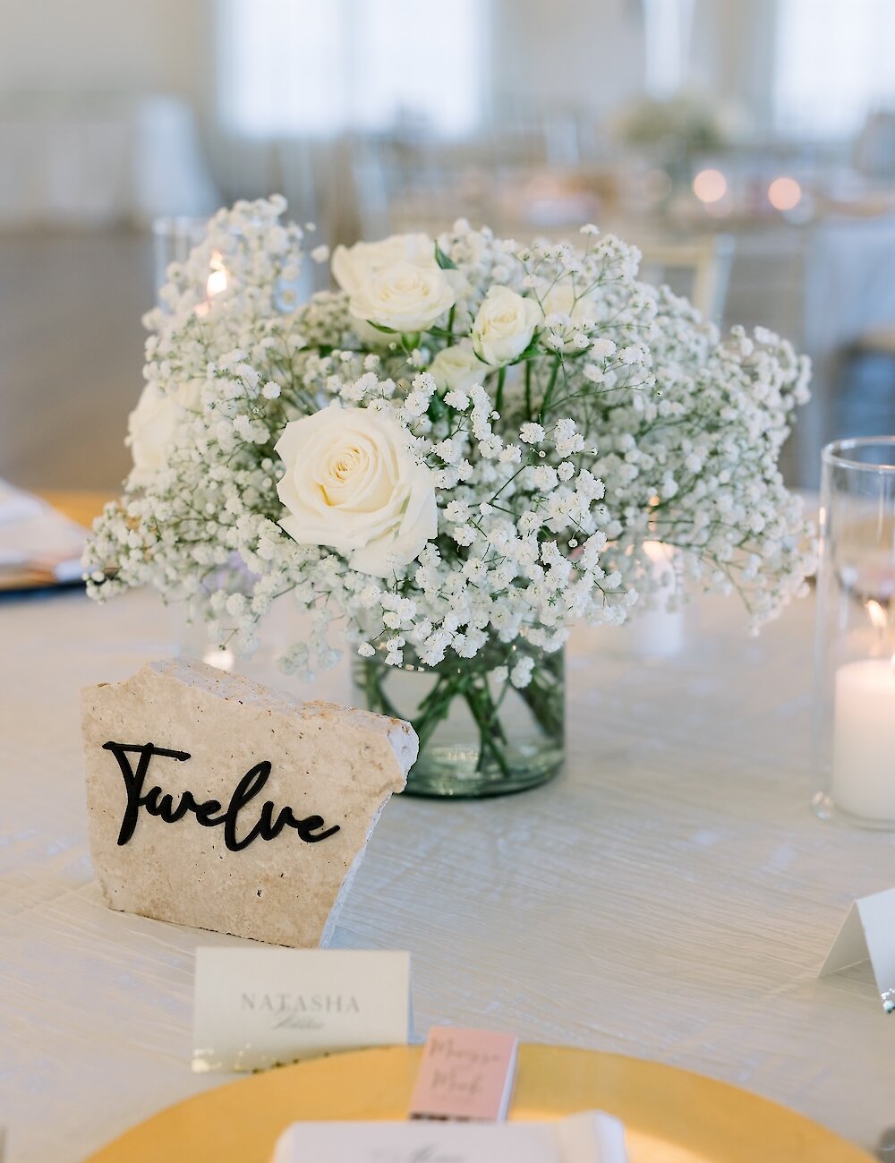A table setting featuring a rustic stone table marker displaying the number 'Twelve' next to a centerpiece of white roses and baby's breath.