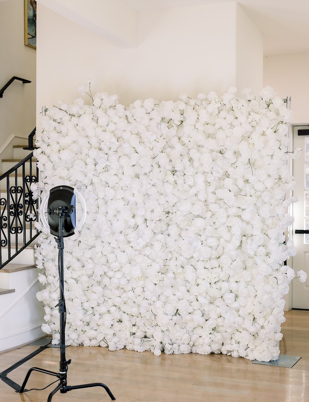 A floor-to-ceiling white flower wall is set up as a photo backdrop with a professional ring light and camera stand next to an elegant staircase.