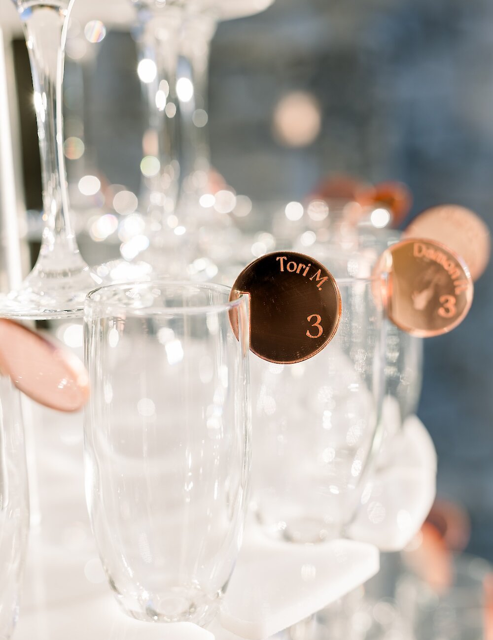 Close-up of champagne flutes on a display tower, each holding a round, copper-colored tag with a guest's name and table number etched on it.