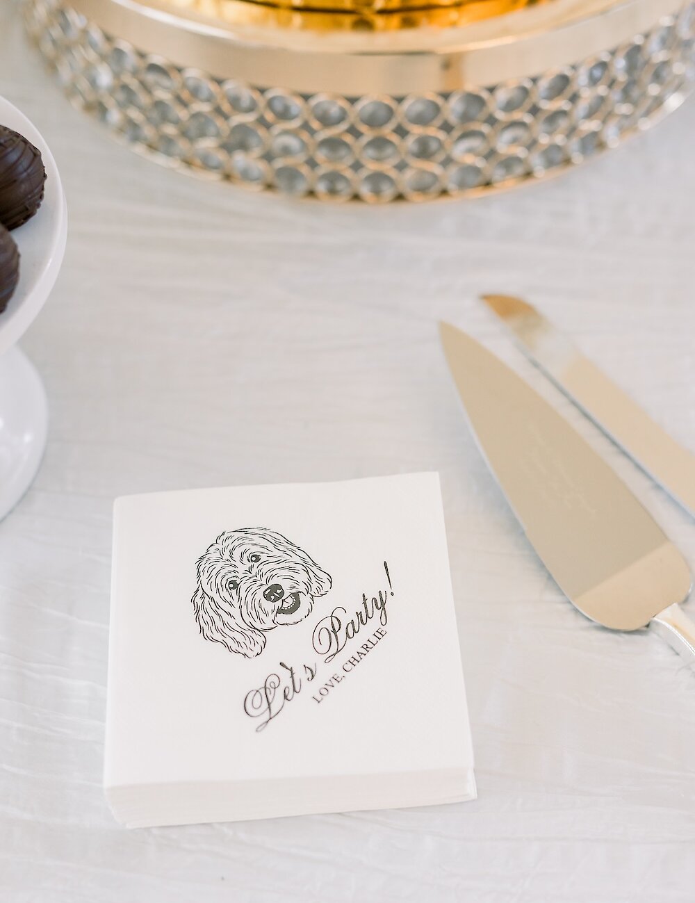 Close-up of a stack of white cocktail napkins personalized with a dog drawing and the text 'Let's Party! Love, Charlie' on the dessert table.