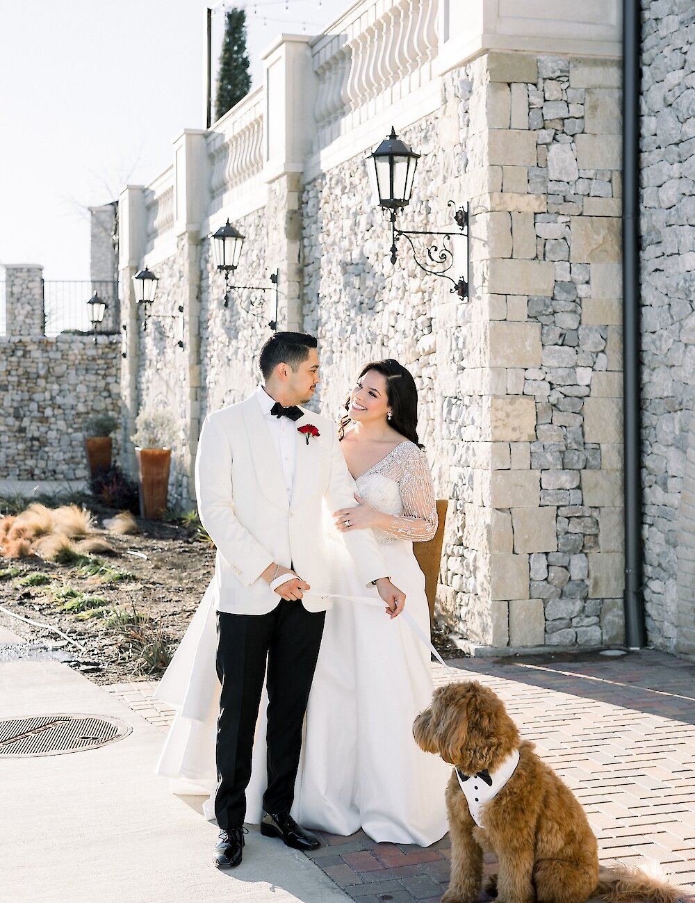 The bride and groom smile at each other while posing with their golden doodle dog, wearing a bow tie, against a white stone wall exterior.