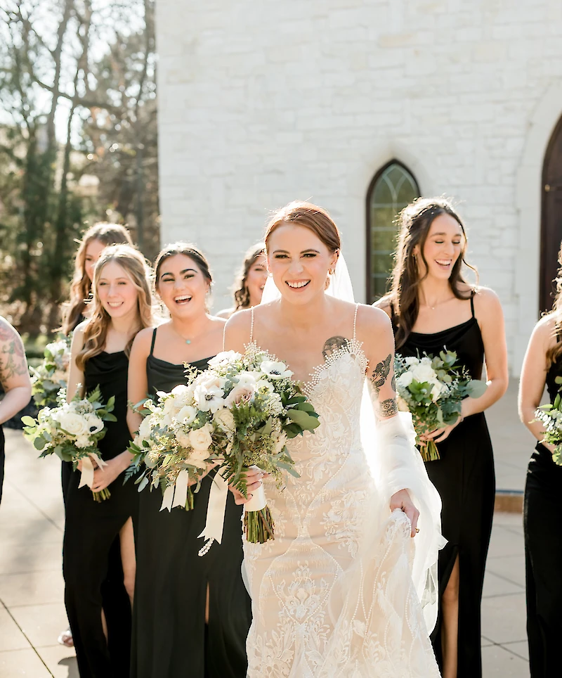 A laughing bride in a white lace gown walks happily, holding a large bouquet, with her bridesmaids in black dresses surrounding her outdoors.