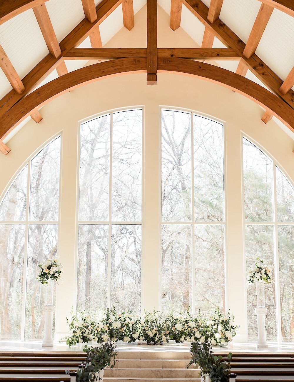 The interior of a bright wedding chapel featuring vaulted wooden beams, large arched windows, and a low altar decorated with lush white florals.