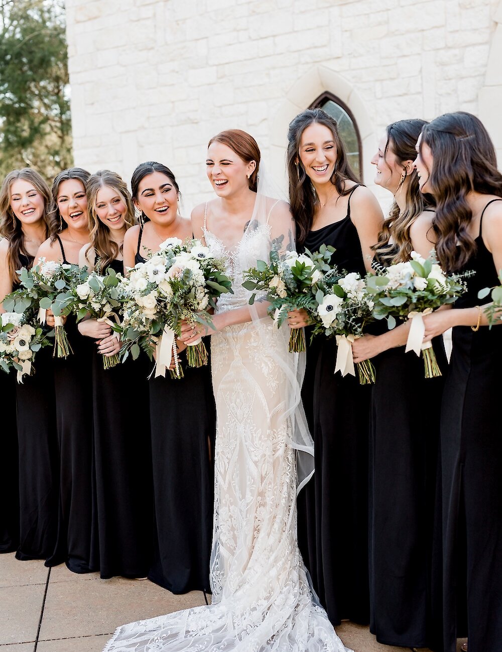 The laughing bride in her white lace gown poses for a photo with her seven bridesmaids, all wearing matching long black dresses and holding bouquets.
