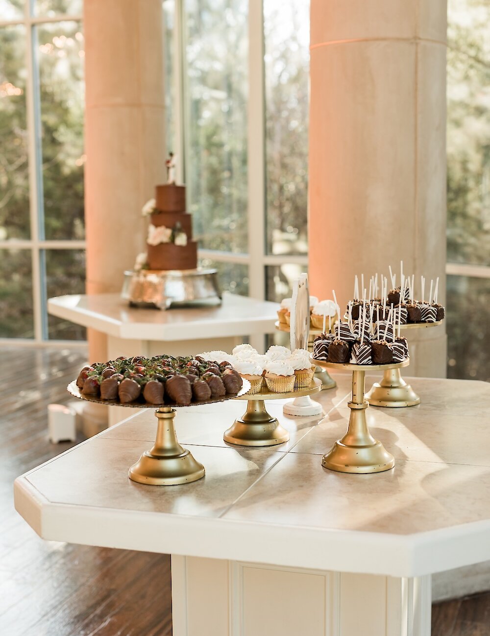 A dessert table featuring tiered platters of chocolate-covered strawberries and cake pops, with a small chocolate wedding cake on a stand in the background.