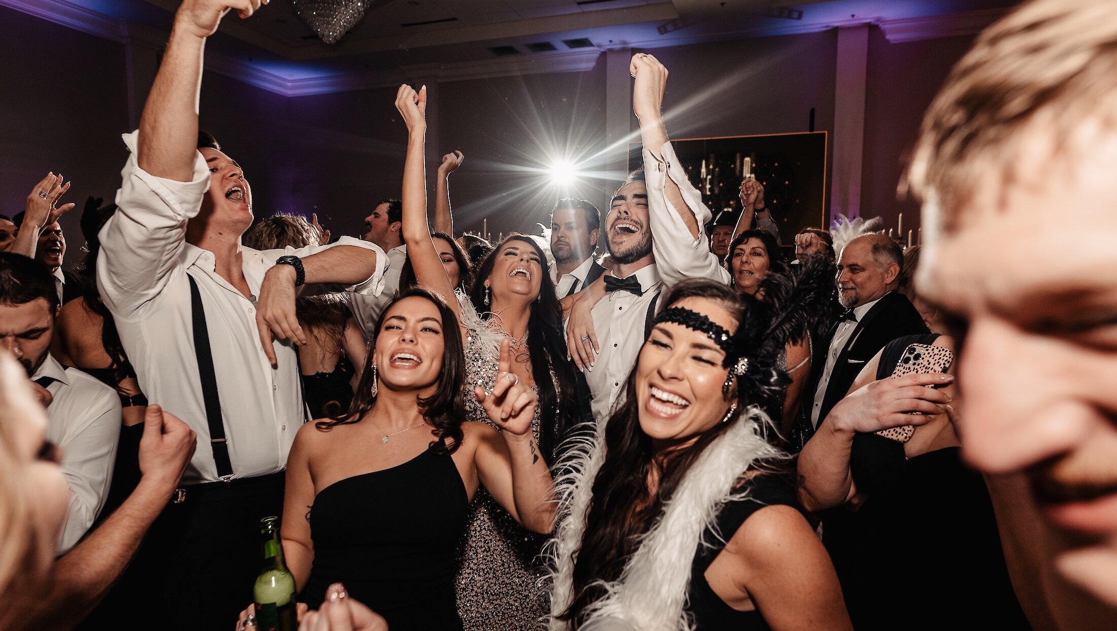 Excited wedding guests, some dressed in Great Gatsby-inspired attire, cheer and dance with their arms raised during a lively reception party.