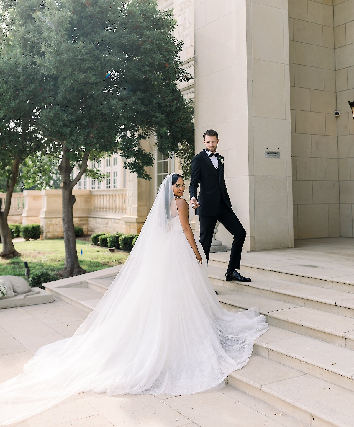 The bride and groom descend grand stone steps outside a luxurious mansion venue, with the bride looking back as her long veil and train flow behind her.