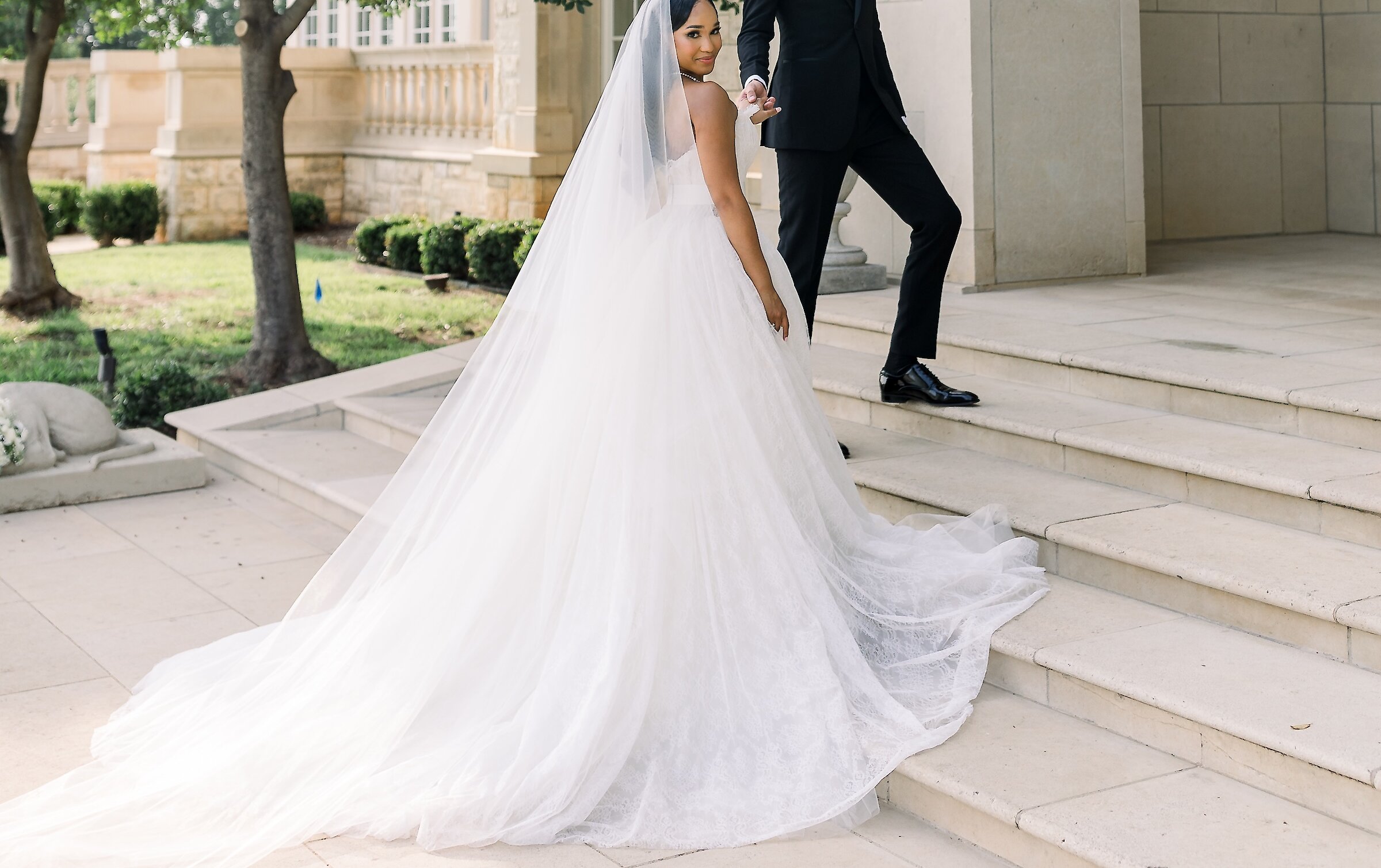 The bride and groom descend grand stone steps outside a luxurious mansion venue, with the bride looking back as her long veil and train flow behind her.