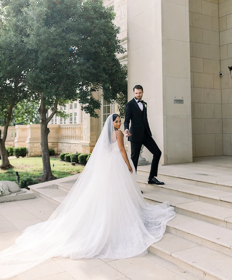 The bride and groom descend grand stone steps outside a luxurious mansion venue, with the bride looking back as her long veil and train flow behind her.