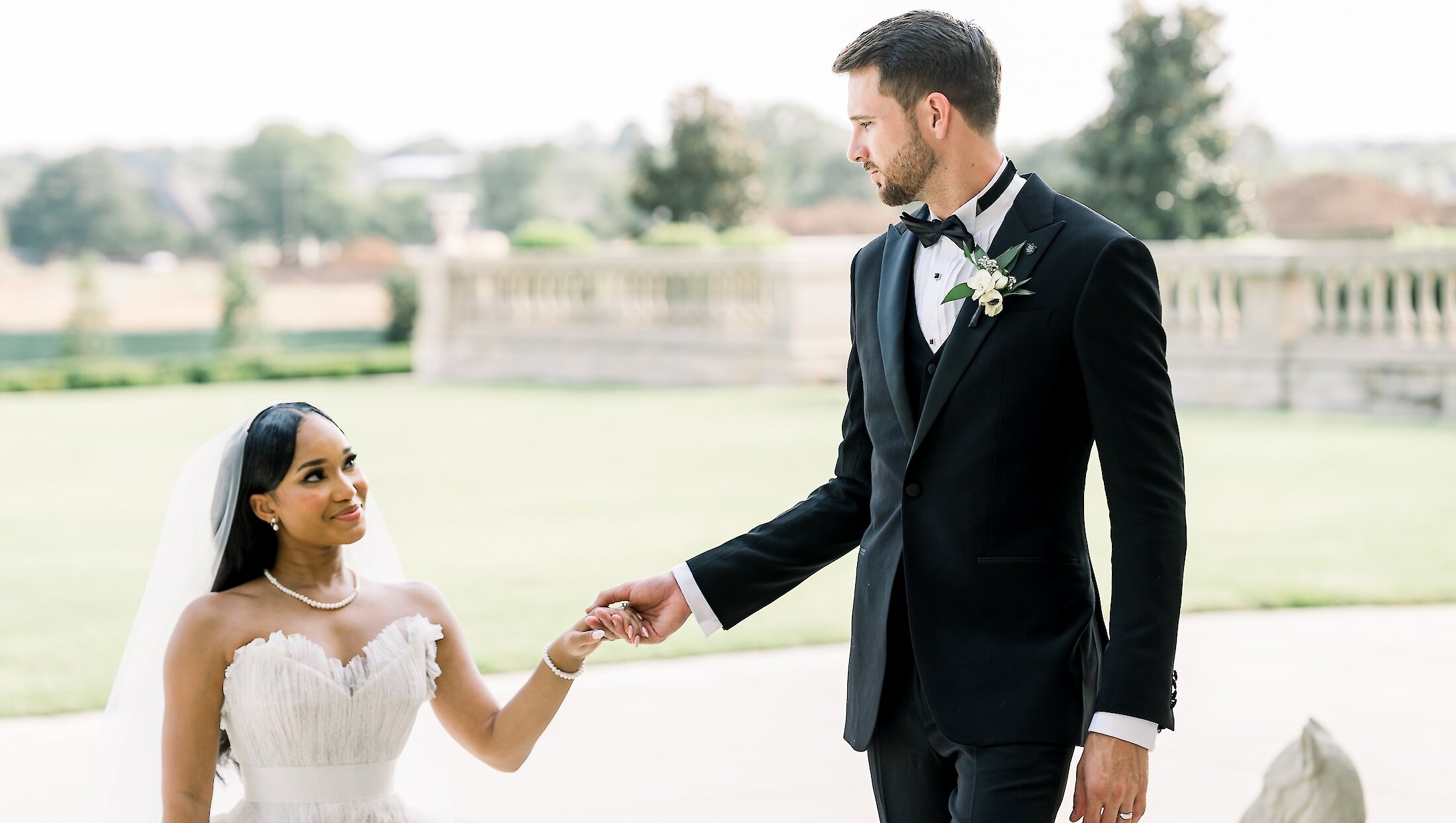 The bride in a ruffled strapless gown holds hands with the groom in a tuxedo, smiling at him while walking across a paved area near a sprawling lawn.